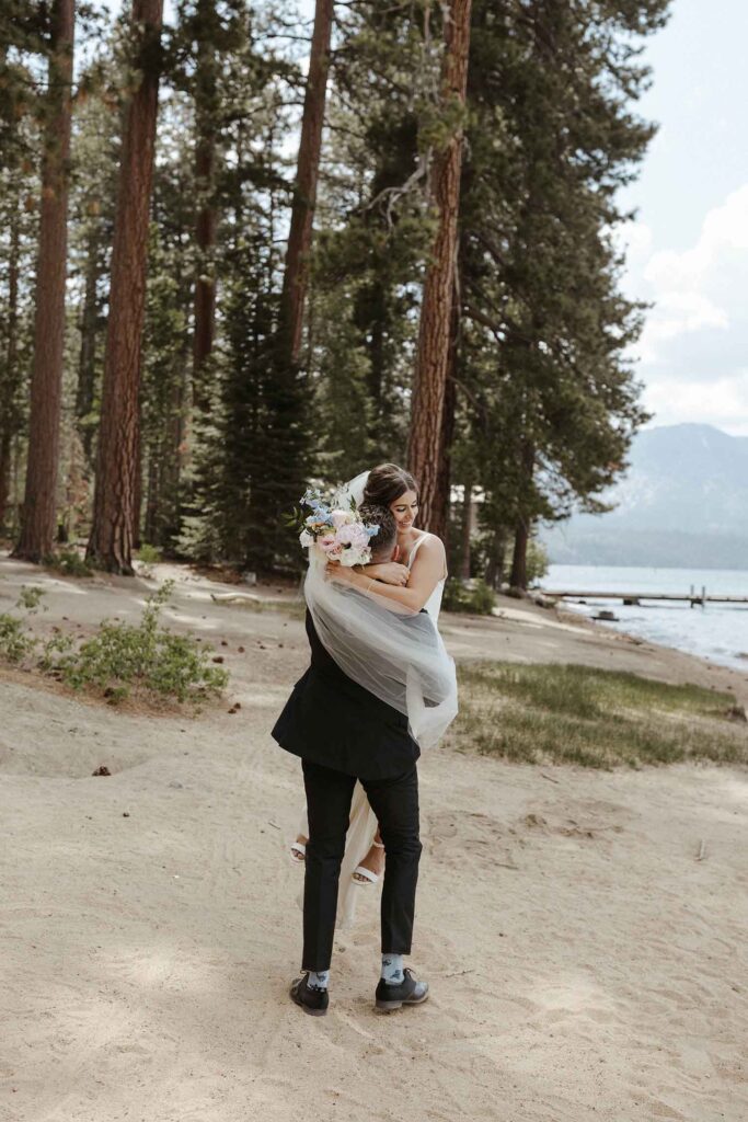Groom lifting elopement bride up in the air and spinning her around in Emerald Bay with tall trees in background