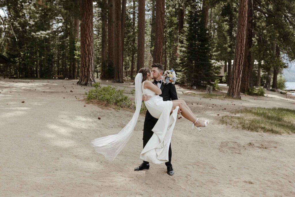 Groom holding bride in the air and kissing her with tall trees in background at Emerald Bay
