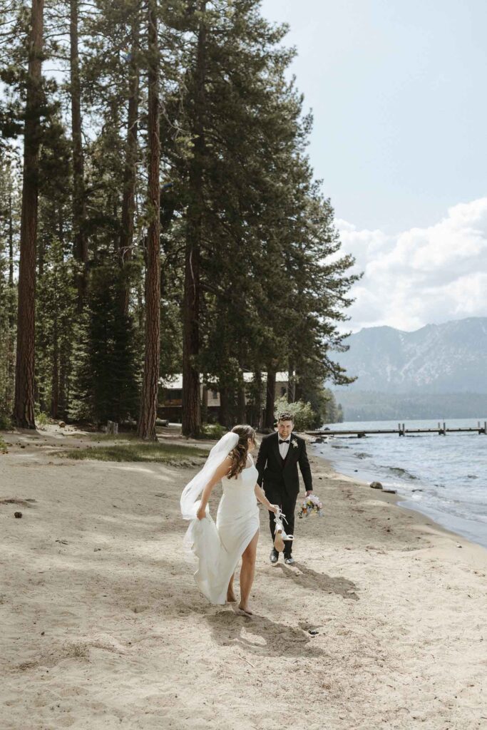 Elopement bride running along sandy beach toward camera while holding wedding shoes in her hand and looking back at groom at Emerald Bay