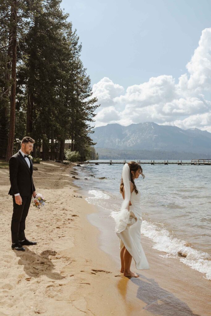 Bride standing with bare feet on wet sand and looking out toward Emerald Bay while groom watches