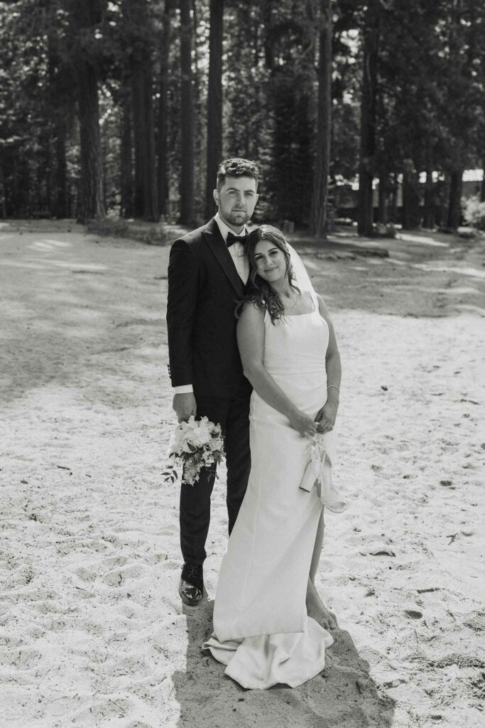 Elopement couple standing together on sandy beach and looking at camera with tall trees in background at Emerald Bay
