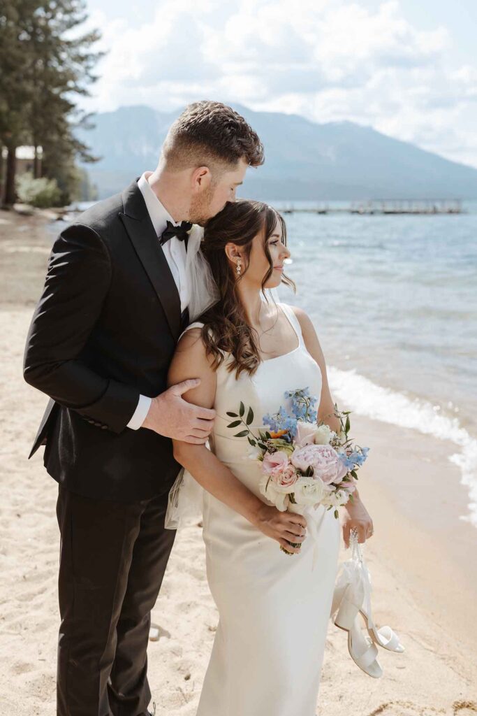 Groom kissing elopement bride on head while holding her arm and she looks out towards Emerald Bay with water and mountains in background