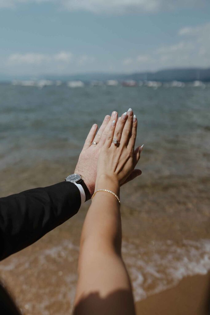 Close up of elopement couple holding hands up next to each other while wearing wedding rings with Emerald Bay in background