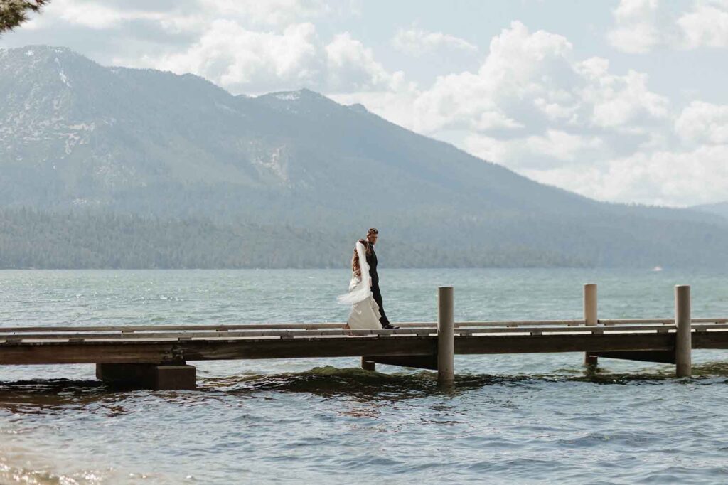Elopement couple holding hands while walking out along pier in water together at Emerald Bay with tall mountains and pine trees in background