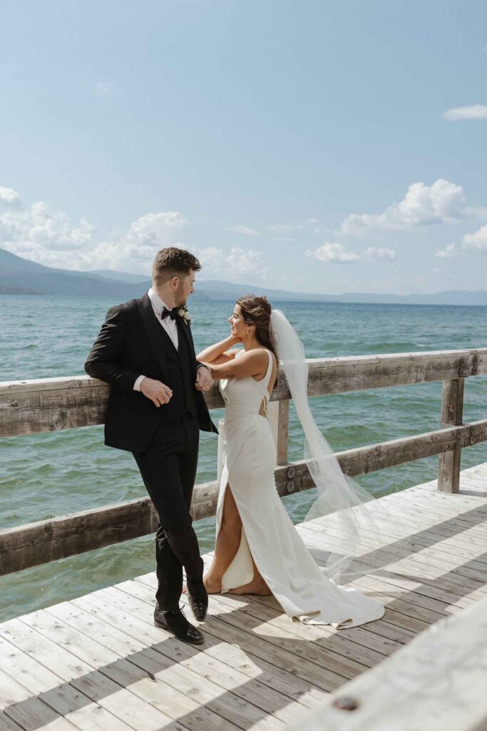 Groom leaning on wooden railing on pier and talking to Bride while she looks out at water at Emerald Bay