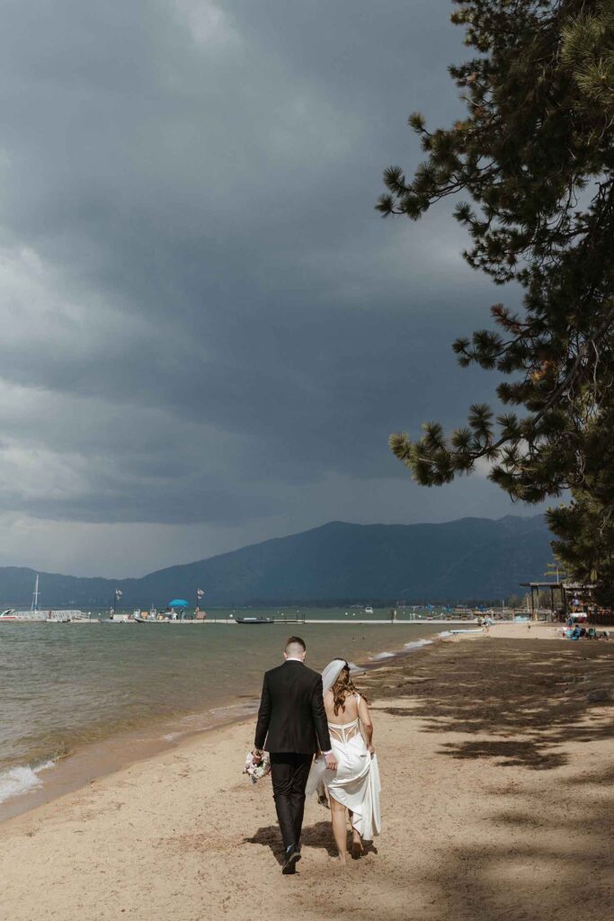 Elopement couple walking along sandy beach together at Emerald Bay next to pine trees while storm clouds loom above