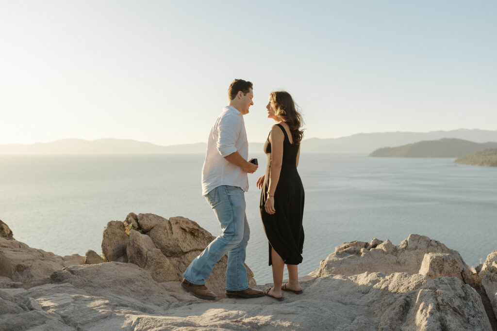 Man smiling at woman as he starts to kneel while standing atop Eagle Rock with Lake Tahoe in background