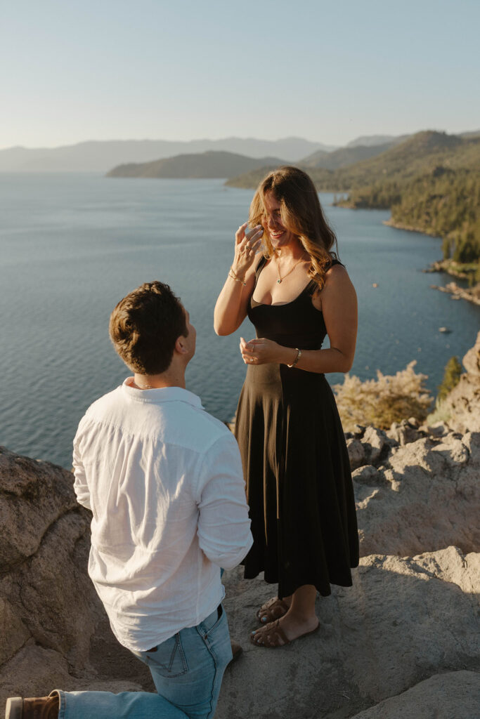 Woman smiling while fixing hair and man is on one knee in front of her at Eagle Rock with Lake Tahoe in background