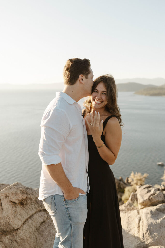 Woman showing off engagement ring and smiling while fiancé kisses head at Eagle Rock in Lake Tahoe