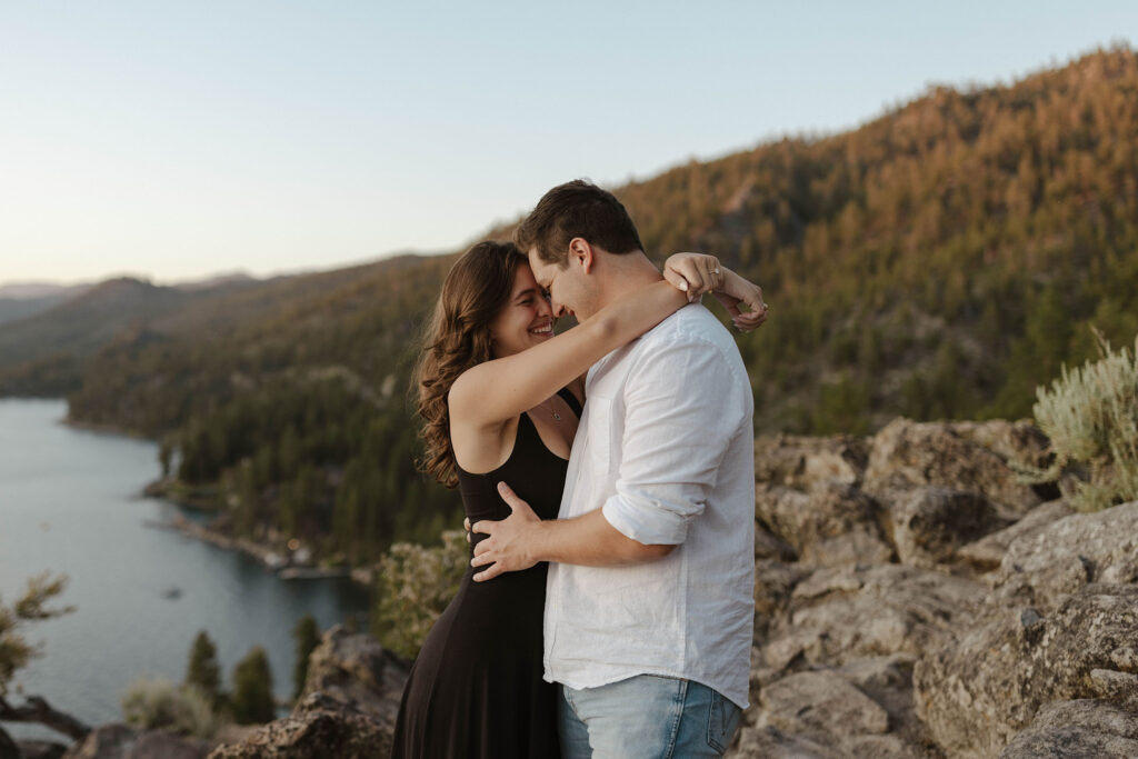 Engagement couple holding and smiling at each other at Eagle Rock with trees and Lake Tahoe in background