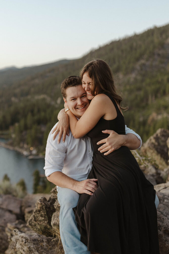 Woman sitting on fiancé's lap while they both hug and smile at Eagle Rock in Lake Tahoe