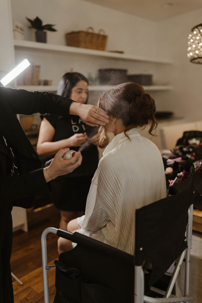 Hair stylist spraying hair spray on brides hair while she sits in a chair during getting ready inside at Nakoma Resort