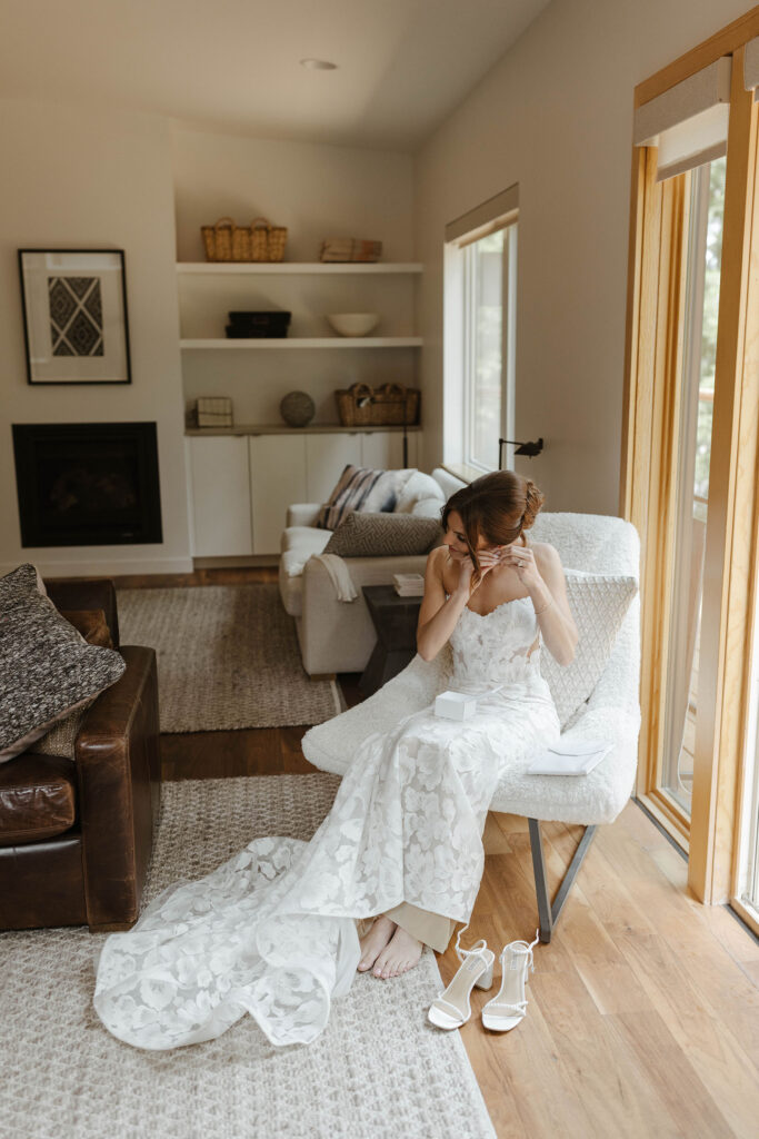 Bride in wedding dress sitting in white chair and putting on earrings inside at Nakoma Resort