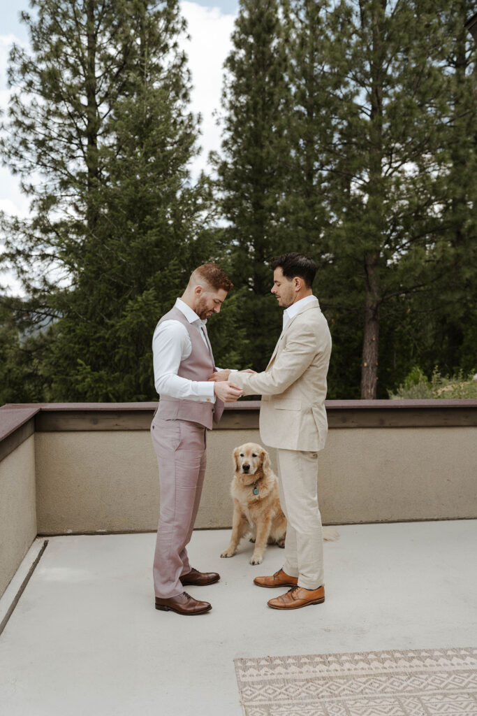 Groomsmen helping wedding groom fasten vest buttons outside while dog watches at Nakoma Resort