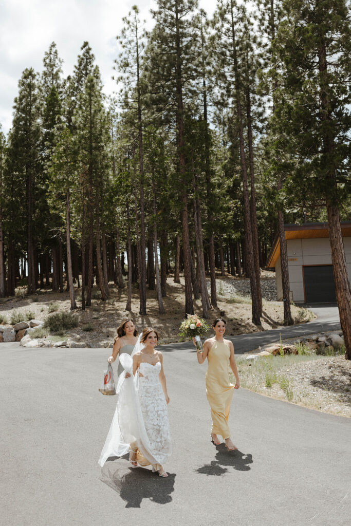 Wedding bride walking down road next to bridesmaids holding floral bouquets with tall pine trees in background at Nakoma Resort