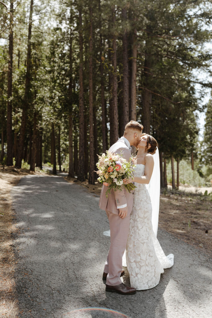 Wedding couple kissing while standing on pathway together and bride holds colorful floral bouquet in front of tall pine trees at Nakoma Resort
