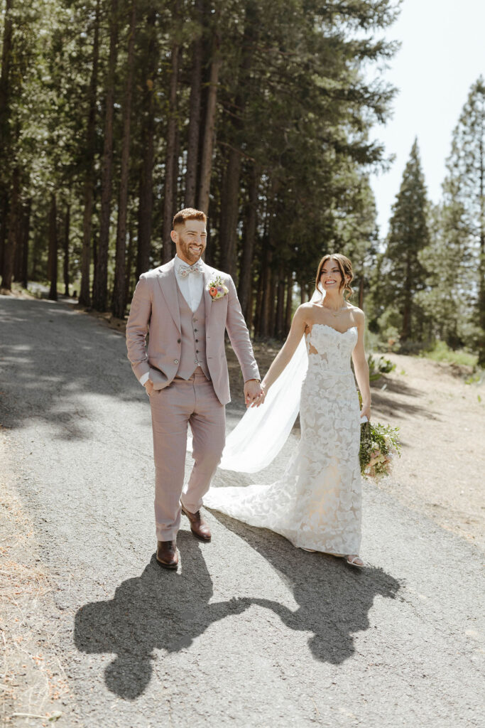 Wedding couple holding hands while walking down roadway and smiling together with tall pine trees in background at Nakoma Resort