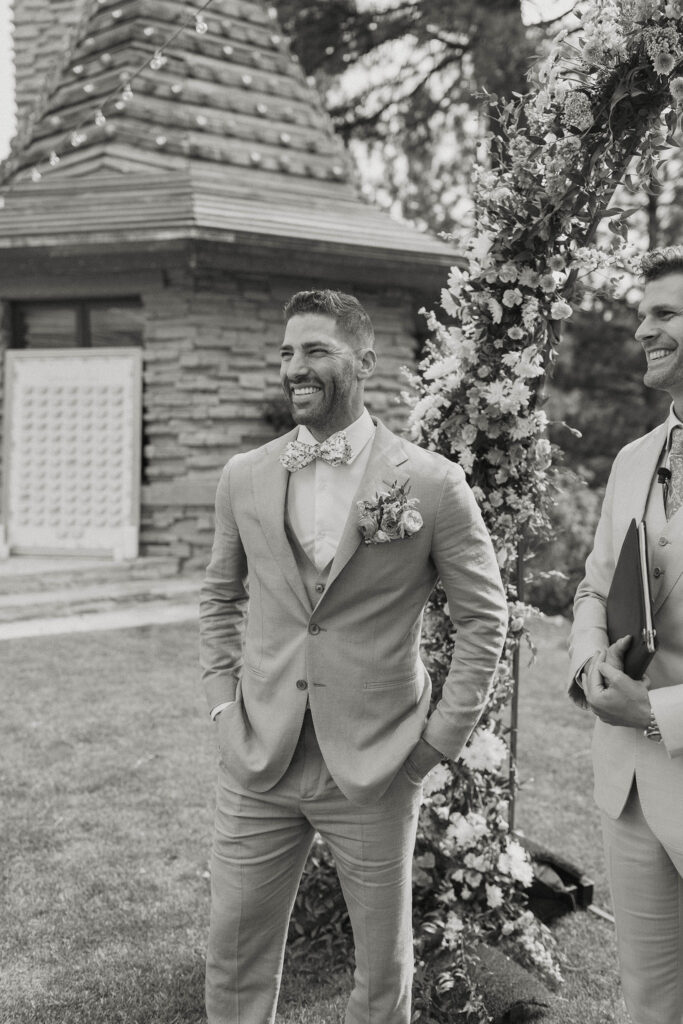 Groom with hands in his pockets and smiling while standing at wedding ceremony watching bride walk down aisle at Nakoma Resort