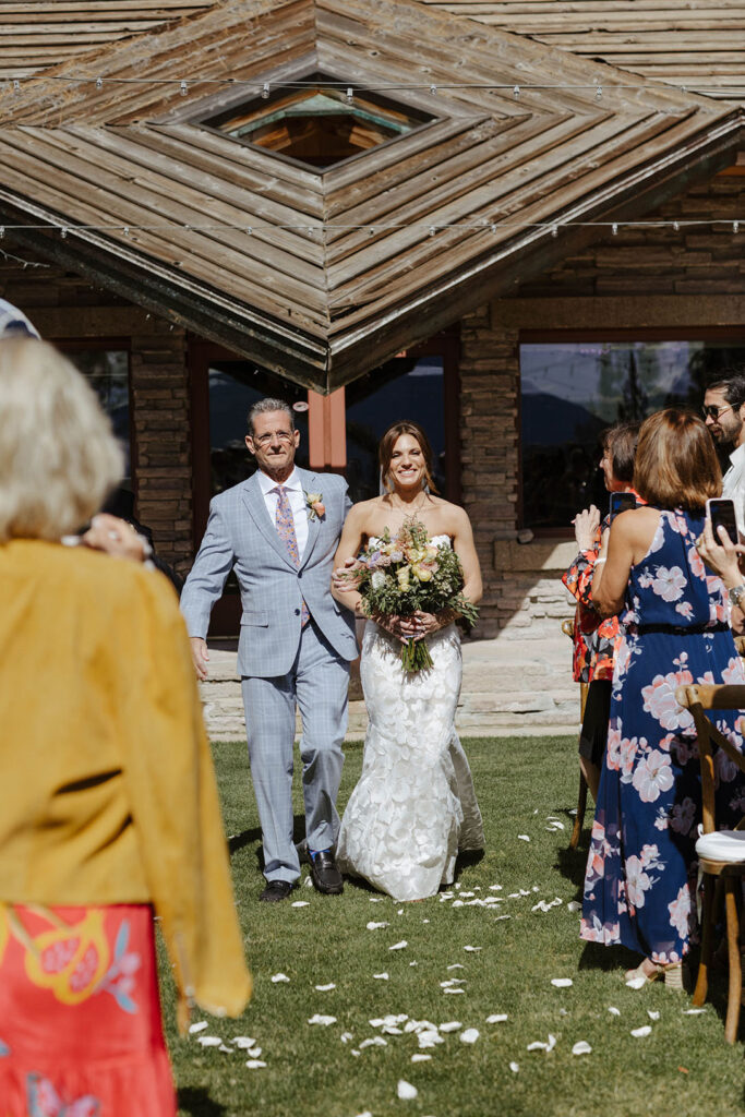 Bride holding floral bouquet while walking down wedding aisle covered in flower petals and dad holds her arm at Nakoma Resort