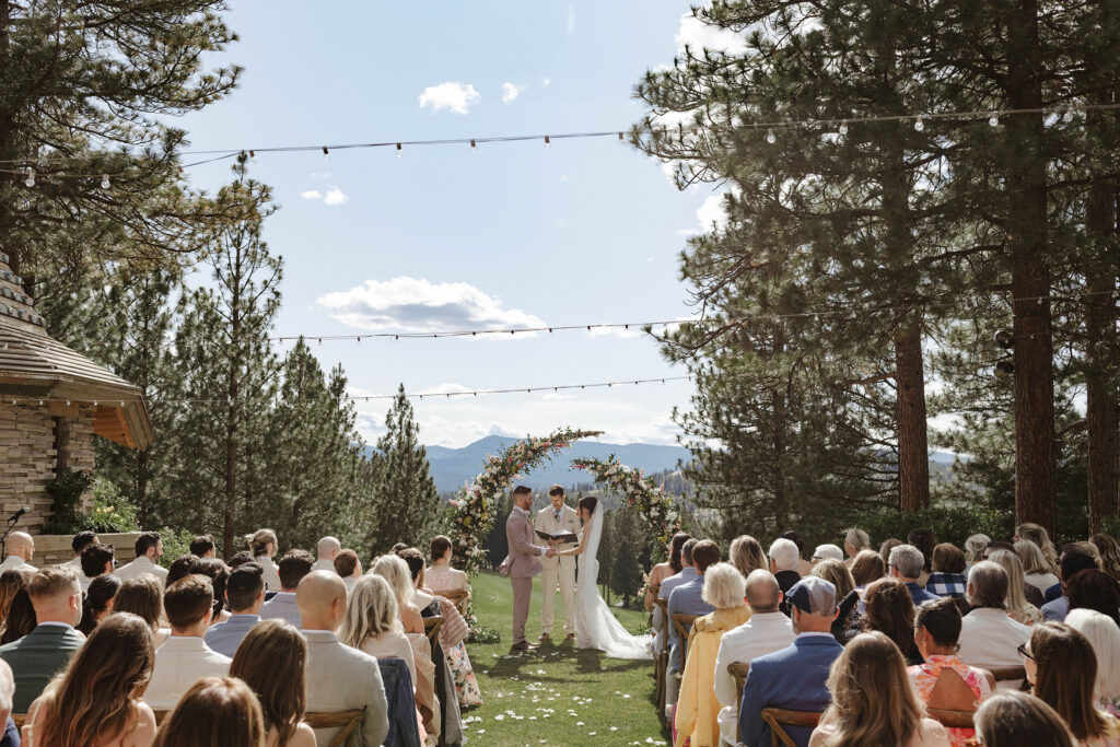 Wedding couple holding hands during ceremony at Nakoma Resort with guests seated and surrounded by tall pine trees