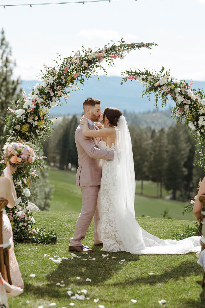 Wedding couple holding each other and kissing in front of arch after ceremony at Nakoma Resort with pine trees and mountains in background