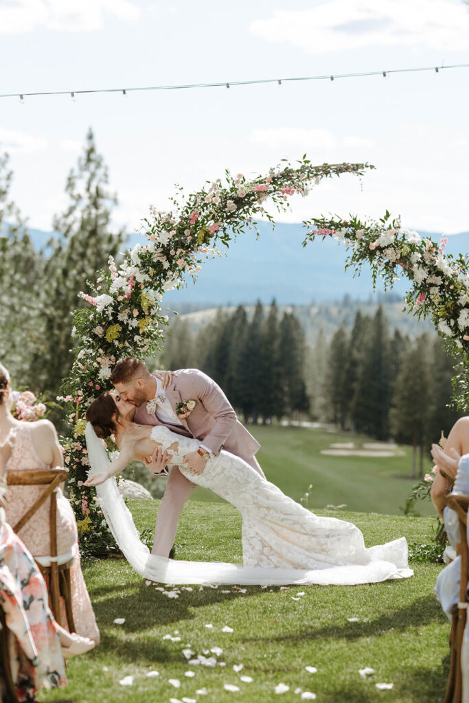 Groom leaning bride back and kissing her in front of wedding arch after ceremony at Nakoma Resort with trees and mountains in background