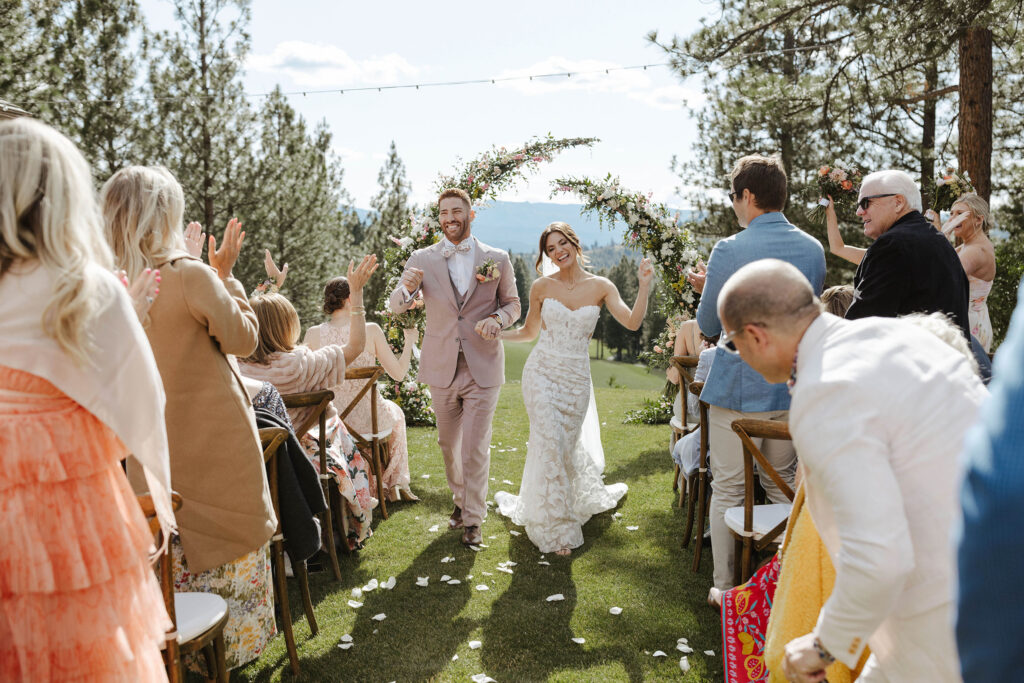 Wedding couple holding hands and smiling while walking down aisle after ceremony and guests cheer at Nakoma Resort