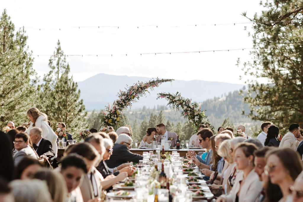 Wedding couple kissing while sitting at head table in front of floral wedding arch during dinner while guests eat around them at Nakoma Resort