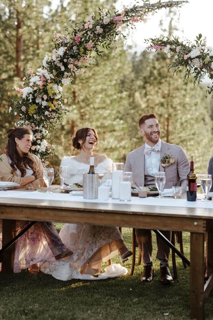 Wedding couple laughing while sitting at wooden table during speeches with floral arch in background at Nakoma Resort