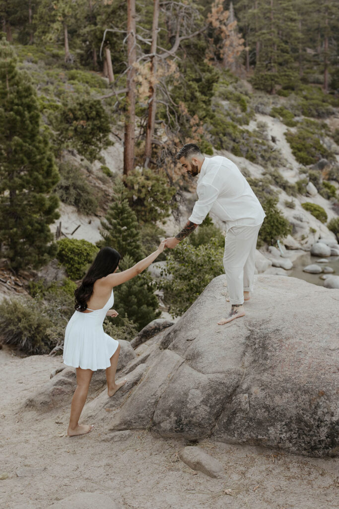 Man standing on large rock helping fiancé up by the hand with trees in background at Lake Tahoe