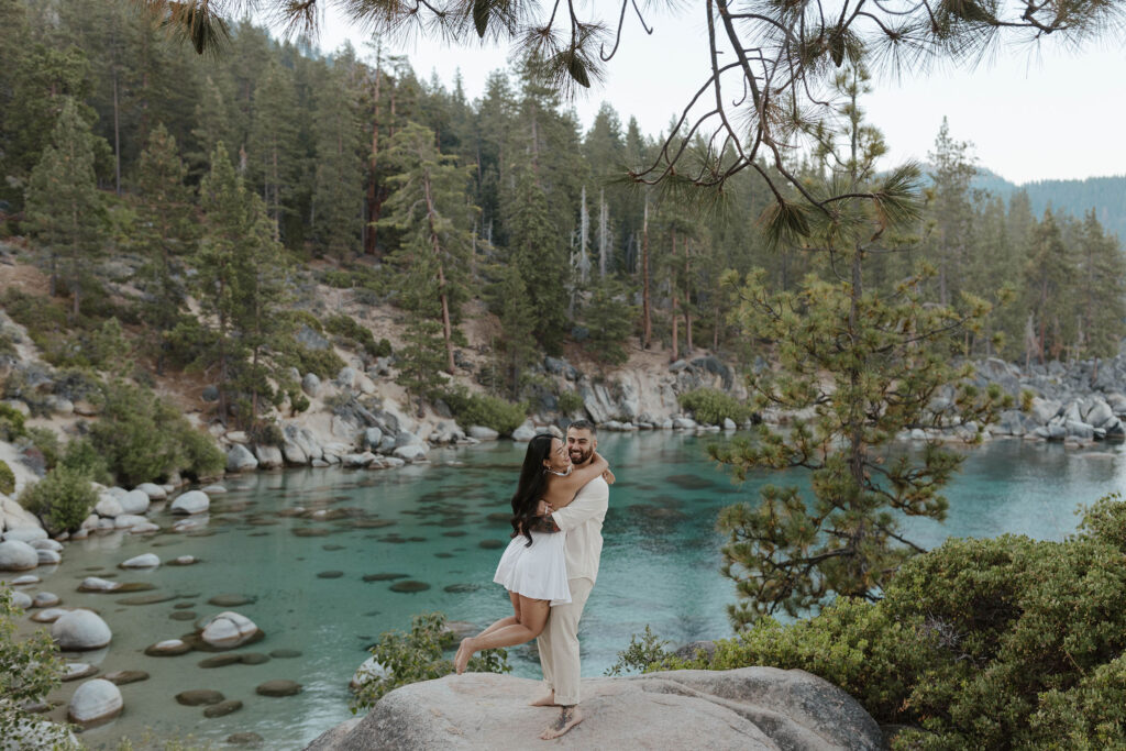 Man picking up fiancé while standing on large rock overlooking Lake Tahoe and pine trees in background