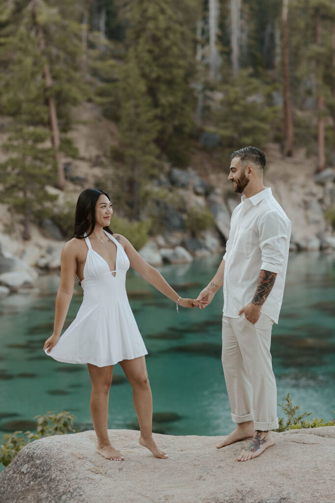 Engagement couple holding hands while smiling at each other and woman plays with dress with Lake Tahoe and trees in background