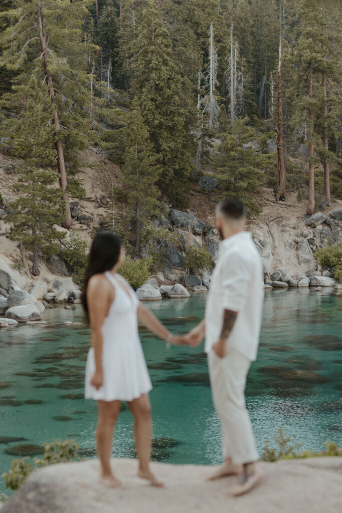 Engagement couple holding hands while standing on rock together and looking out over water at Lake Tahoe