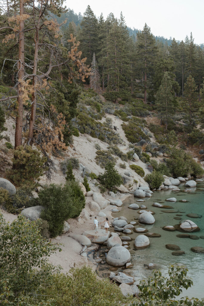 Engagement couple far off in distance walking along large rocks in Lake Tahoe next to beach