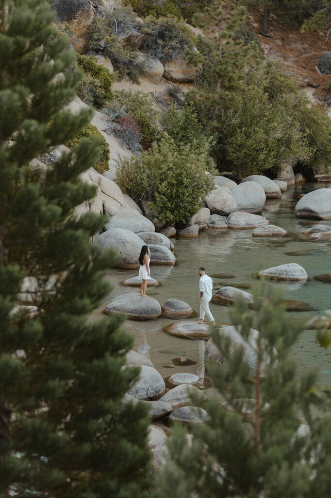 Engagement couple in distance between pine trees walking along large rocks in water in Lake Tahoe