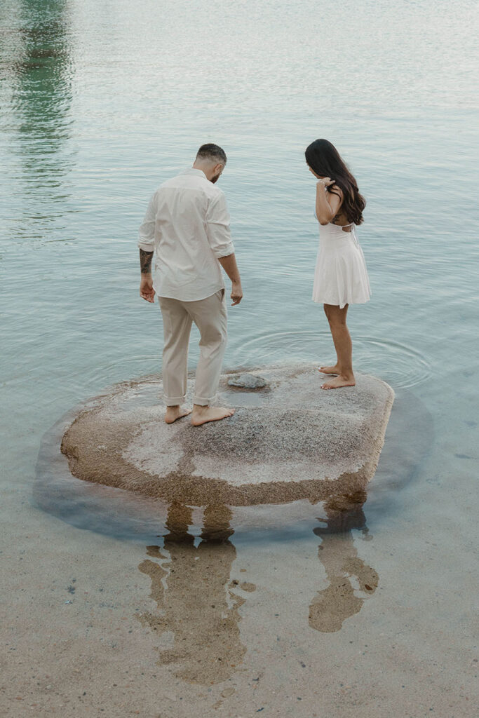 Engagement couple standing on large rock together surrounded by water in Lake Tahoe