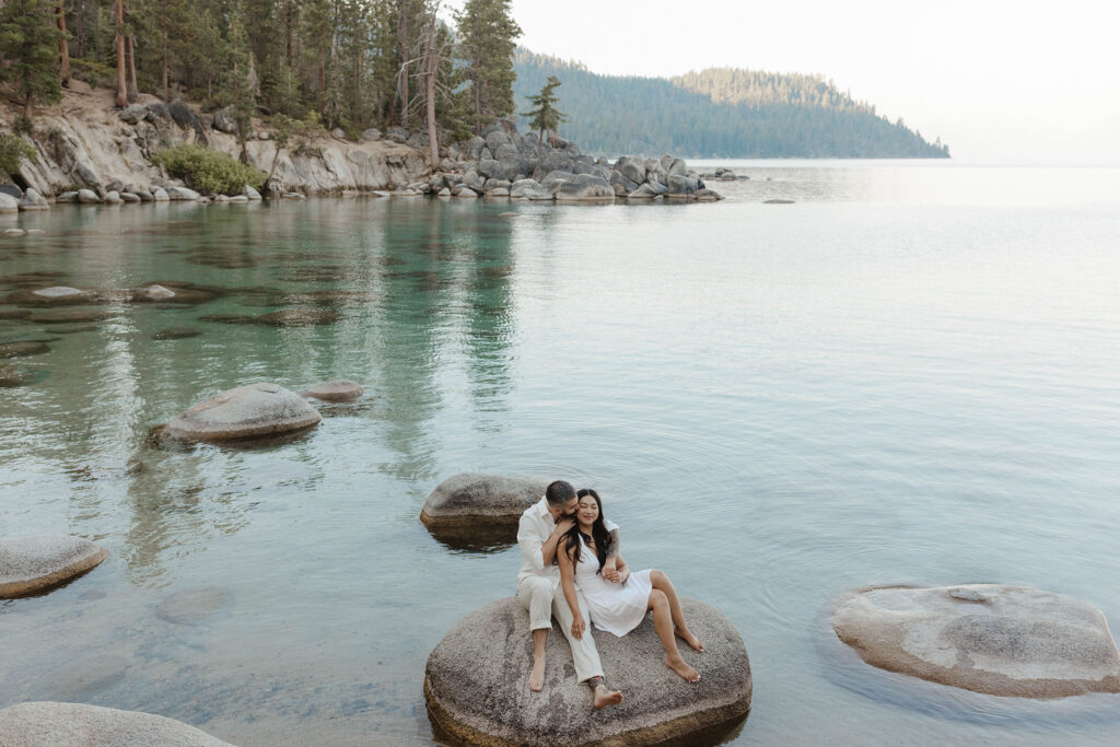 Engagement couple sitting on large rock together surrounded by water while man kisses fiancé on cheek with Lake Tahoe in background