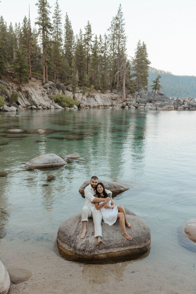 Engagement couple sitting on a large rock together and hugging each other while surrounded by water at Lake Tahoe with pine trees in background