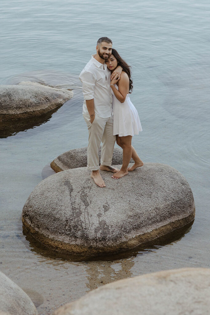 Engagement couple standing on rock in water together while hugging and looking at camera in Lake Tahoe