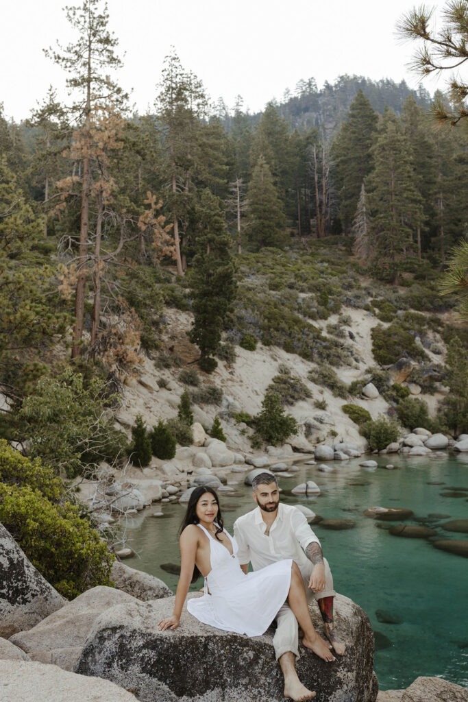 Engagement couple leaning back on large rock together above water with Lake Tahoe and pine trees in background
