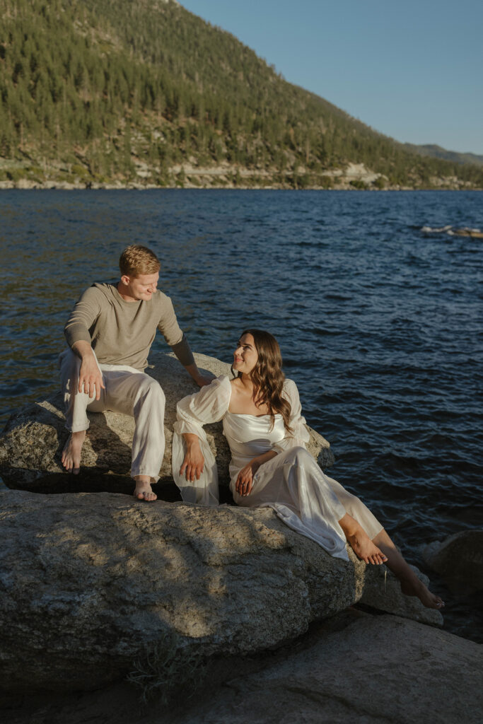 engagement couple sitting on rocks at sunset in lake tahoe looking at each other