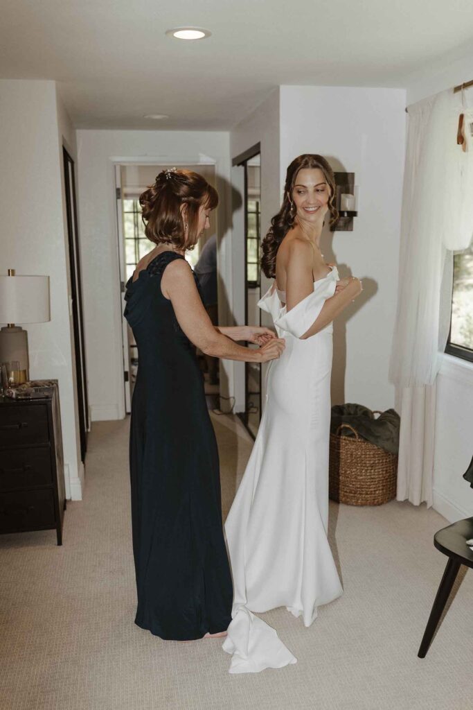 Mom helping elopement bride button back of wedding dress while bride looks over shoulder and smiles in South Lake Tahoe