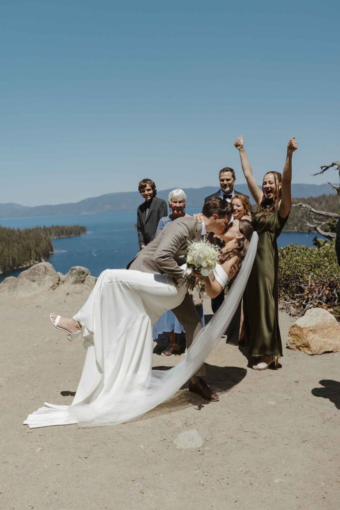 Wedding groom leaning bride back and kissing her after elopement ceremony while guests celebrate in background in South Lake Tahoe