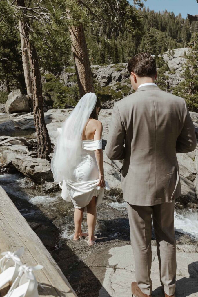Bride holding dress up while walking across stream and groom stands behind her in South Lake Tahoe