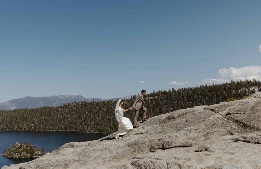 Groom holding bride's hand while leading her up rocky hill in South Lake Tahoe with pine trees and Emerald Bay in background