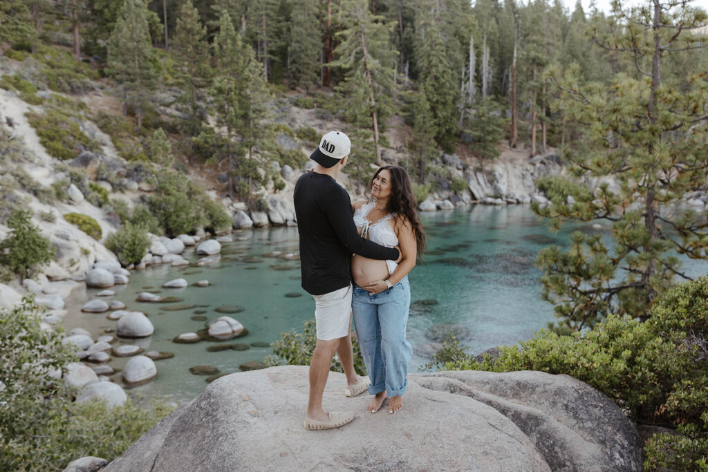 Married couple standing on large rock together overlooking beach in Lake Tahoe while wife holds pregnant belly and husband is wearing Dad hat
