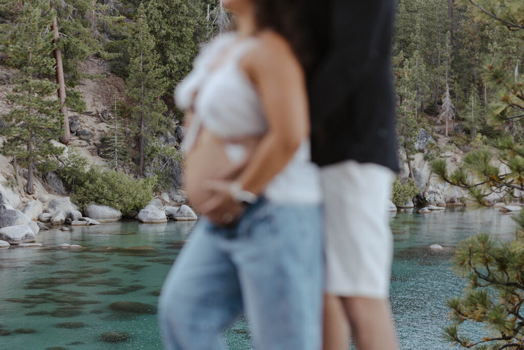 Close up of husband hugging wife from behind while both hold pregnant belly with rocks, pine trees and Lake Tahoe in background
