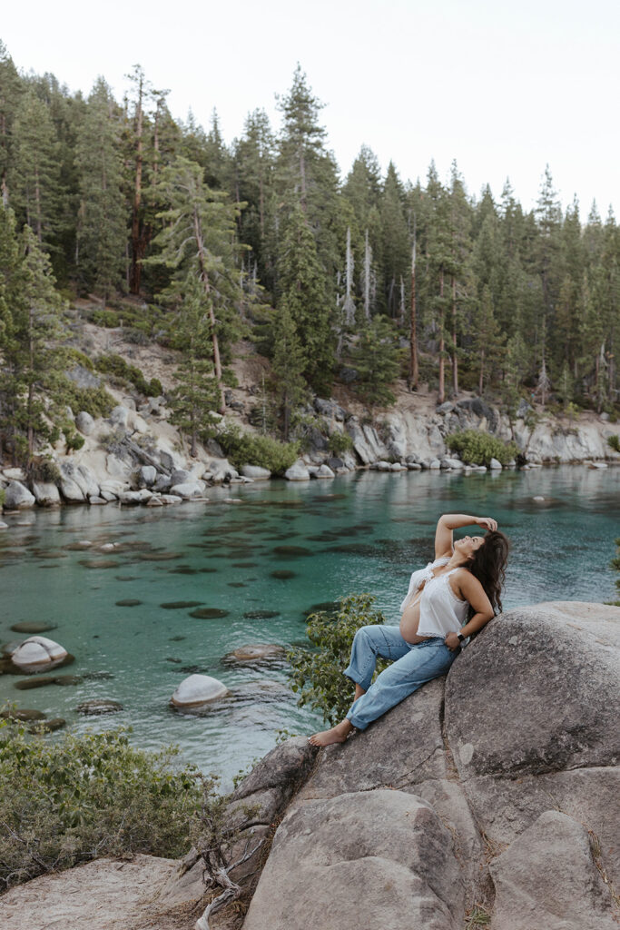 Woman leaning back on large boulder and playing with her hair while showing off pregnant belly with Lake Tahoe and pine trees in background