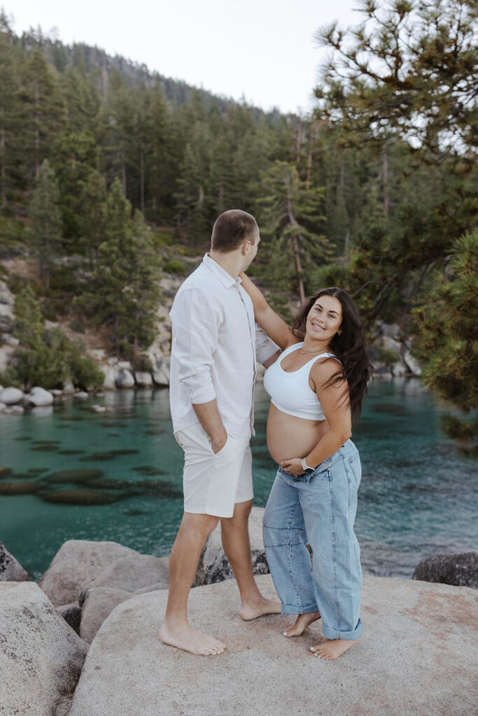 Wife holding pregnant belly and arm on husband's shoulder while he looks out at Lake Tahoe