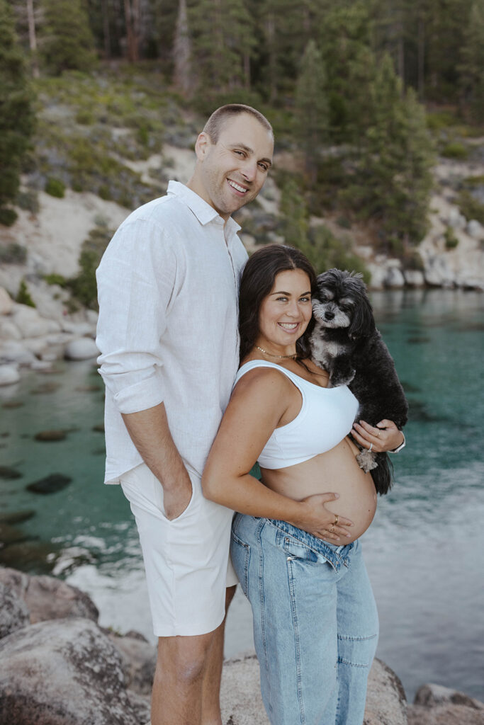 Wife holding dog and her pregnant belly while standing next to husband and smiling at camera in Lake Tahoe